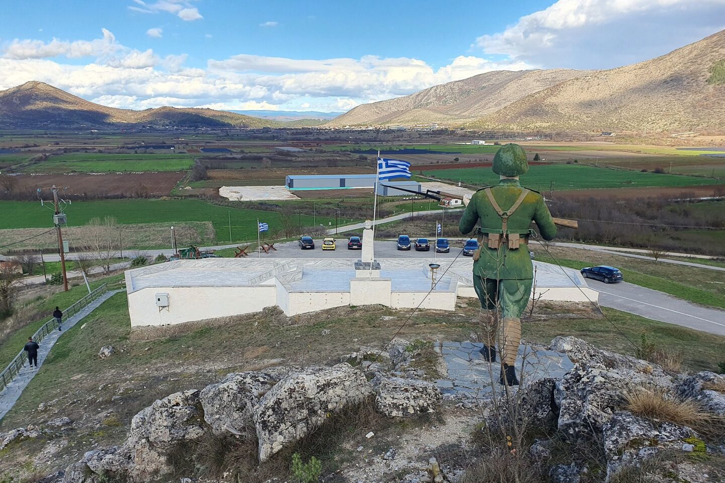 Άγαλμα στρατιώτη με στολή παρατηρεί κοιλάδα δίπλα σε ελληνική σημαία